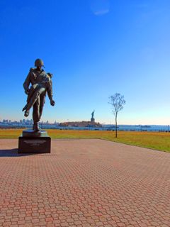 The Statue of Liberty on Liberty Island, viewed from Liberty State Park, the former site of Black Tom Island. In the foreground on the left, <em>Liberation</em>, a bronze statue designed by Nathan Rapoport, depicts a World War II soldier carrying a survivor from a German concentration camp.
